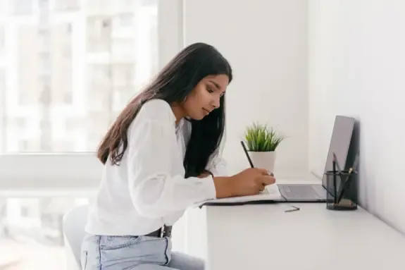 Blog owner writing notes at a bright desk in front of a laptop